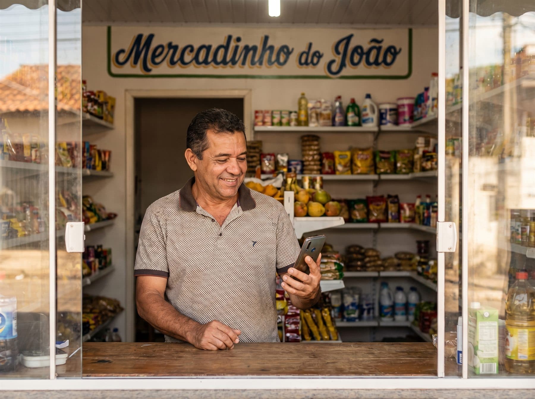 Lojista do Mercadinho do João sorrindo com o celular na mão, atrás do balcão